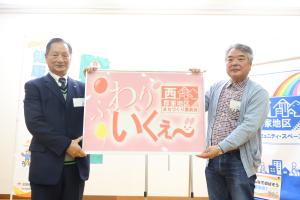 「ふわり いくぇ〜」と書かれた看板を手に持つ老人クラブ会長と西地区郡家地区まちづくり委員会の委員長